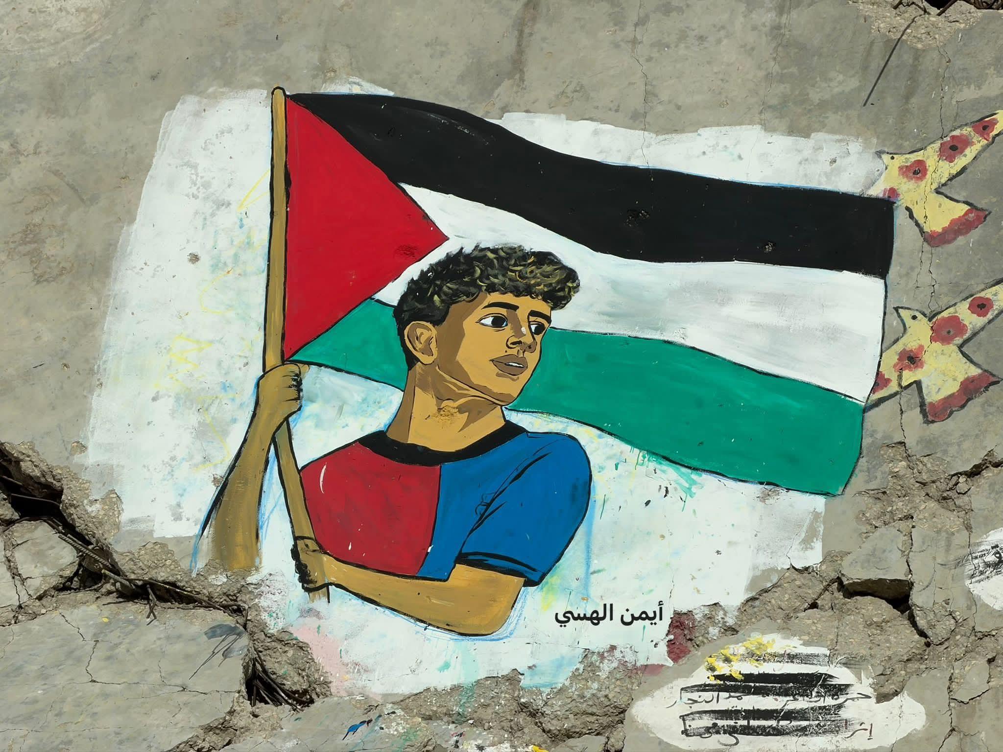 Mural on a damaged wall of a young man holding the Palestinian flag, with yellow doves painted beside it.