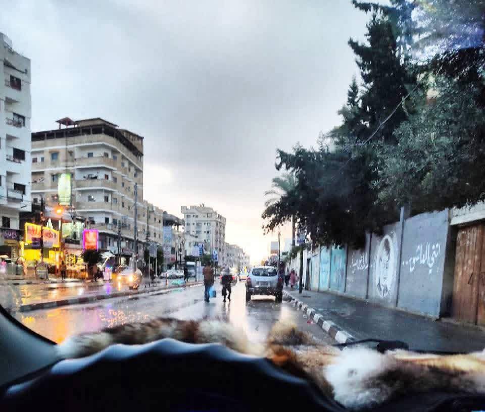 View through a car windshield of a wet city street in Gaza at dusk, with shop lights reflecting on the road.